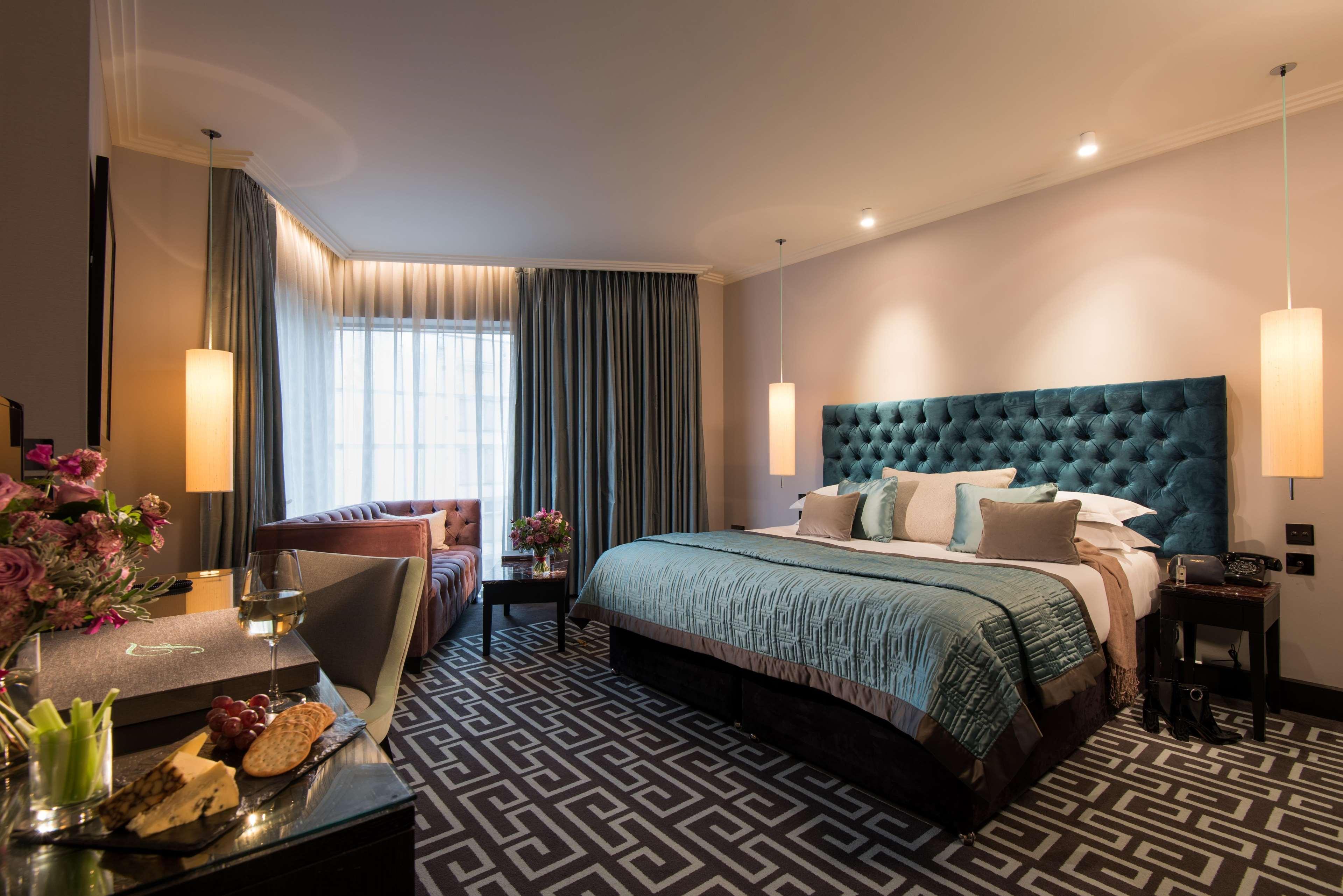 The Fitzwilliam Hotel Белфаст Екстер'єр фото The photo depicts a stylish hotel room. It features a large bed with a tufted teal headboard, flanked by two wall sconces providing soft lighting. The bedding is neatly arranged with accent pillows. To one side, there is a small seating area with a c
