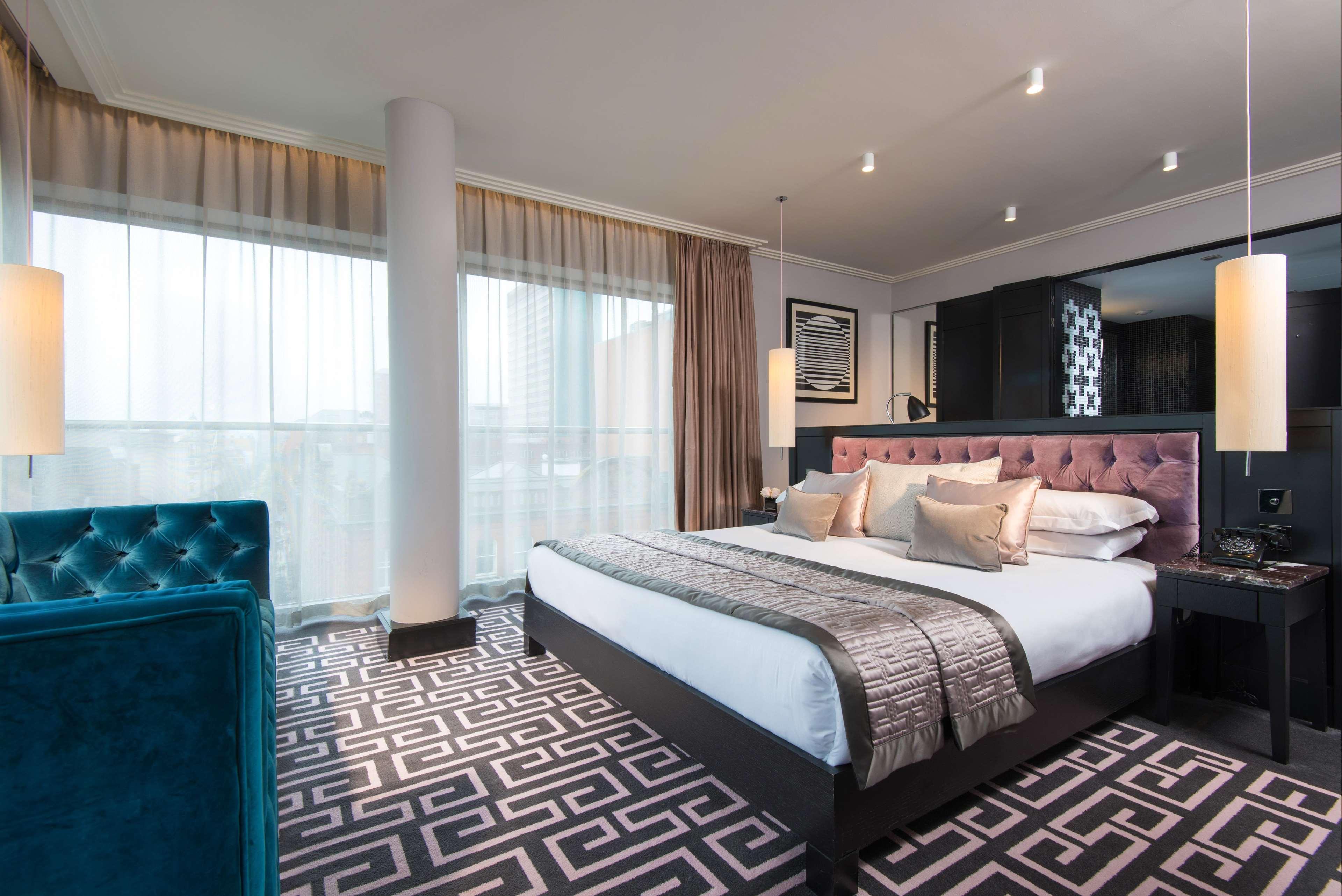 The Fitzwilliam Hotel Белфаст Екстер'єр фото The photo showcases a modern hotel room. It features a large bed with plush pillows and a stylish headboard. The room has large windows with sheer curtains, allowing natural light to filter in. The floor is covered with a patterned carpet that contra